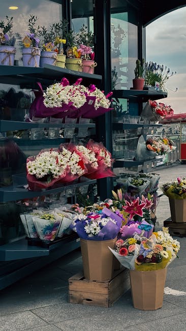 A display of various floral arrangements on black metal shelving and wooden crates outside a florist shop. The arrangement on the middle shelf features white and purple hydrangeas wrapped in bright pi