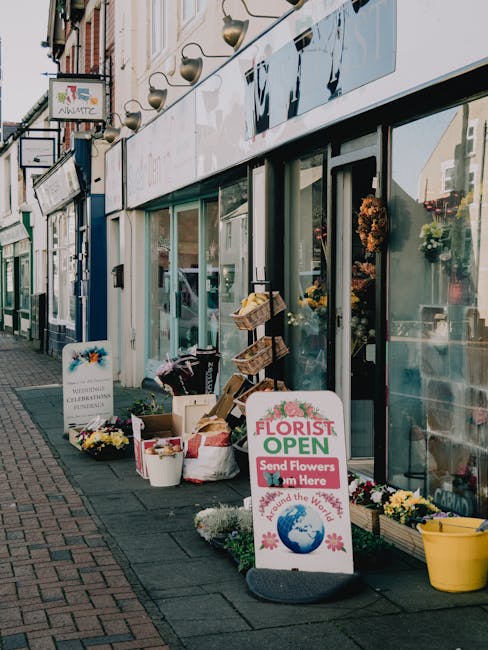 The storefront of Abbey Wood Flowers, a florist shop with large glass windows displaying floral arrangements inside. Outside on the sidewalk, there are various flower displays, including potted plants