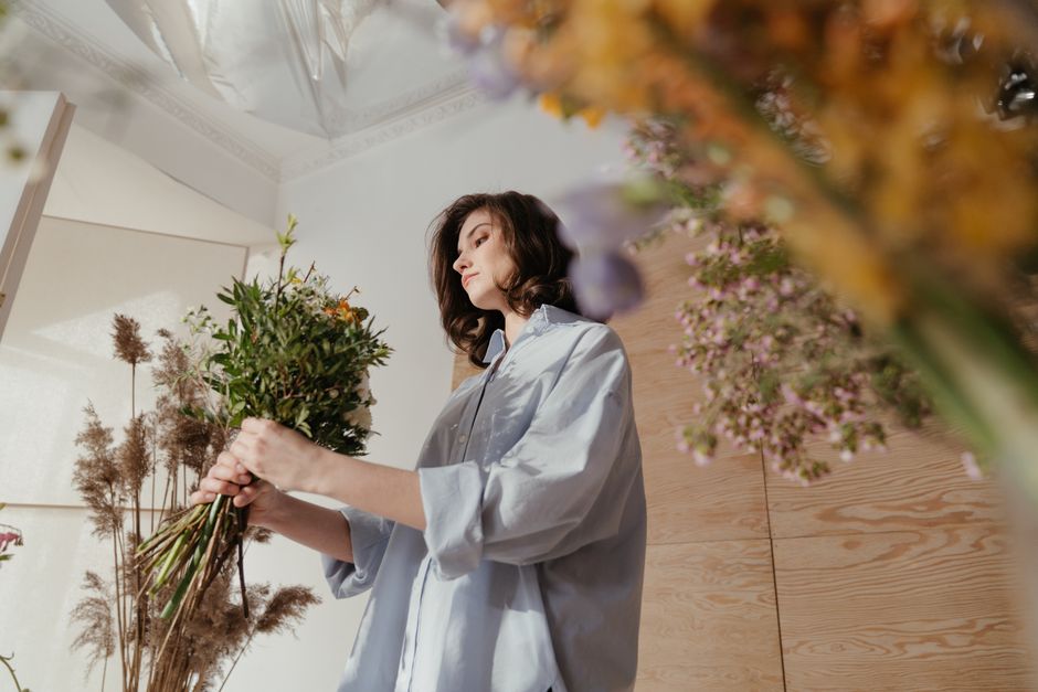 A woman with shoulder-length dark hair, dressed in a light blue shirt with rolled-up sleeves, is holding a bouquet of mixed fresh flowers and greenery. The bouquet includes various flowers and stems, 